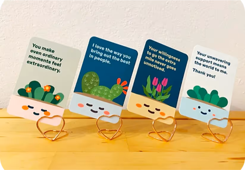 Four Flourish compliment cards with plant illustrations and positive messages, displayed in heart-shaped rose-gold stands on a wooden surface.